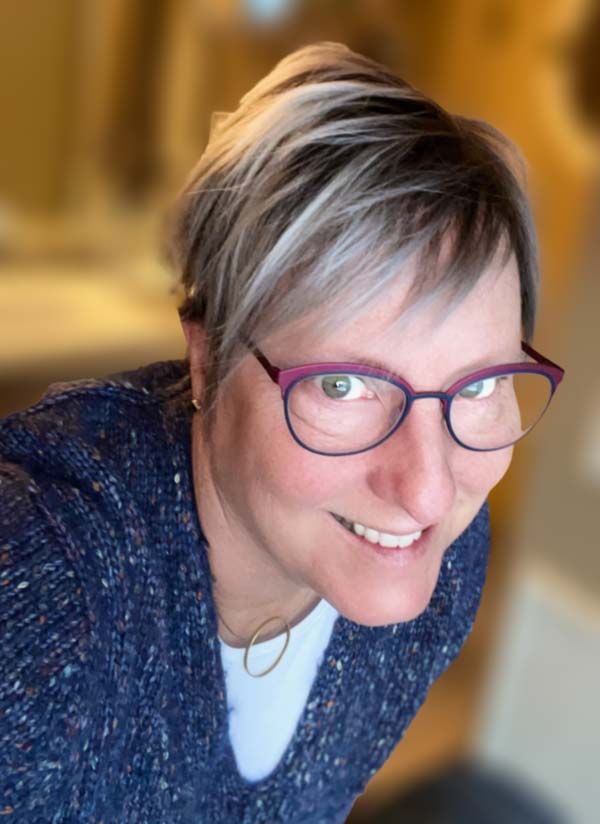 Portrait of Larisa in a blue sweater and glasses smiling at you.